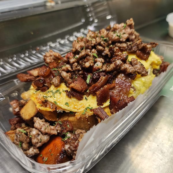 A breakfast bowl with scrambled eggs, potatoes, sausage, and bacon in a clear plastic container.